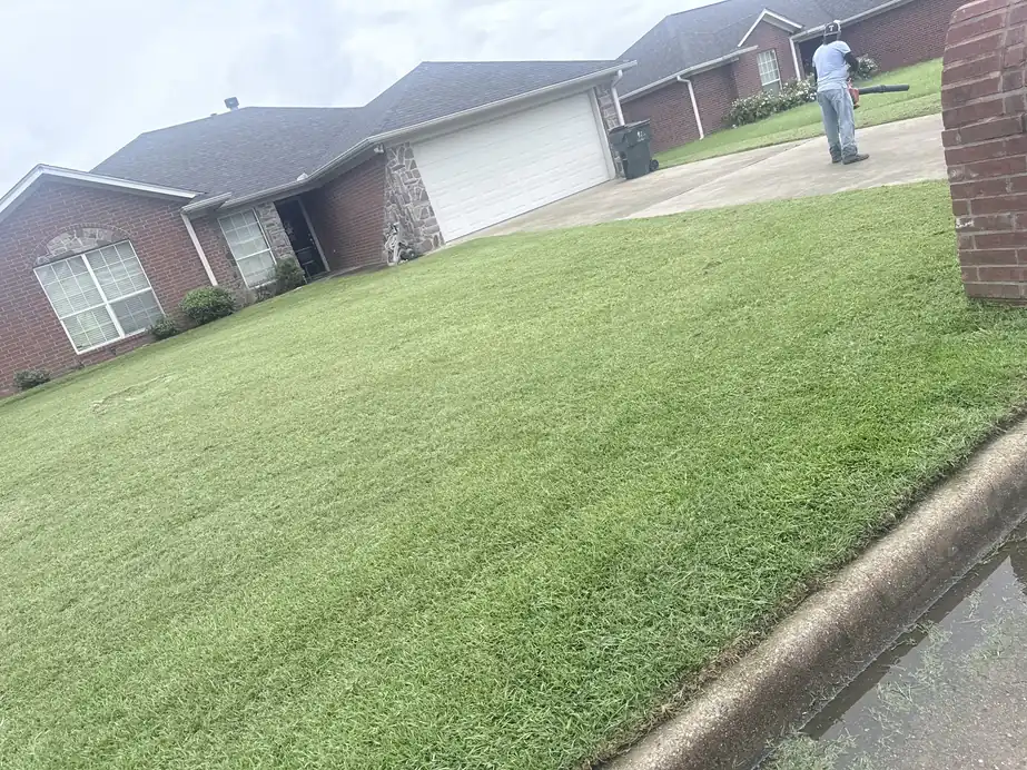 Crew member with a backpack blower clearing a driveway beside a freshly mowed front lawn